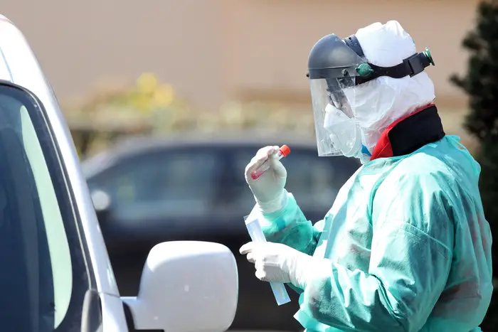 A person in PPE holds a swab by a car at a drive in testing site